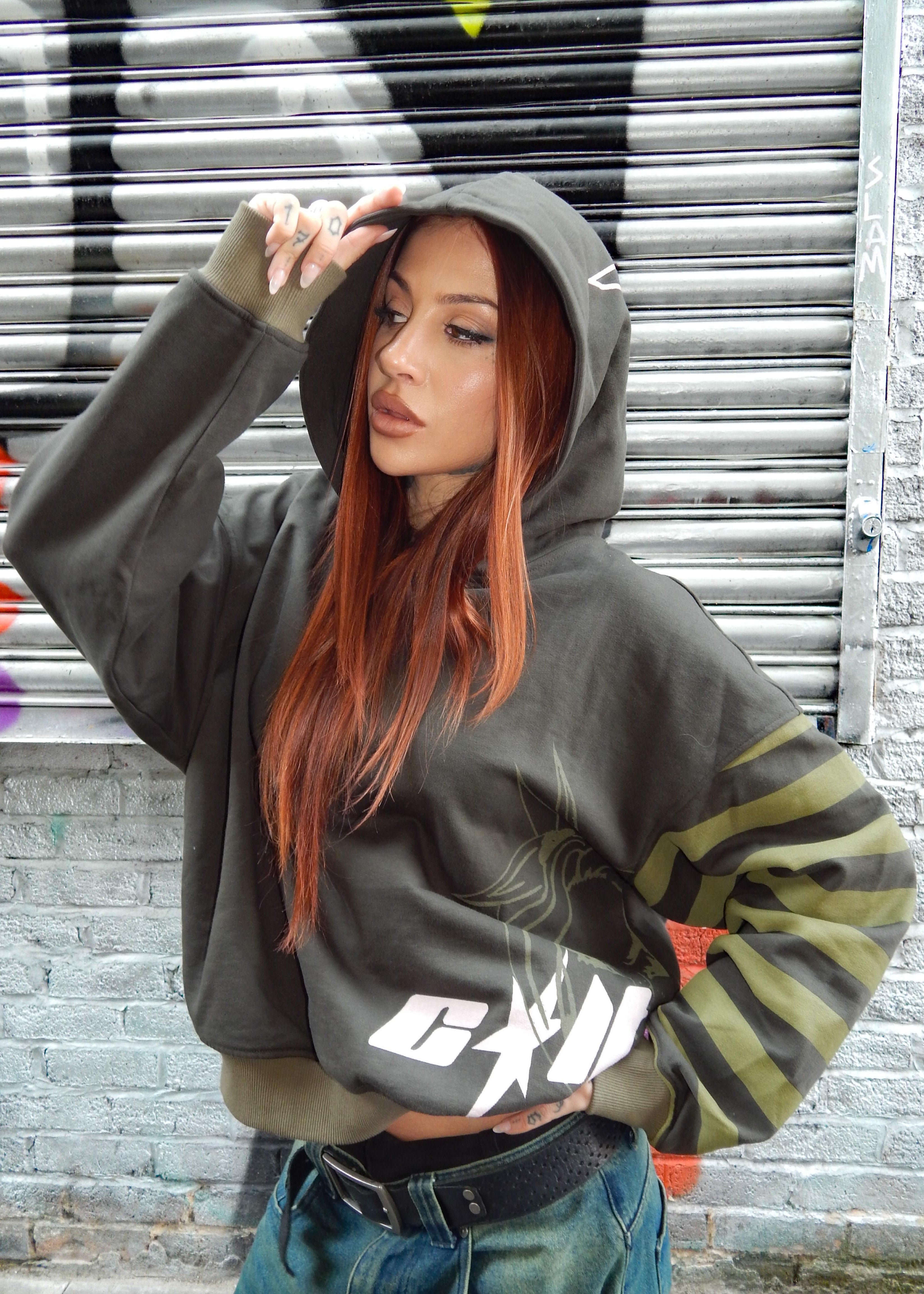 A woman with long red hair models a dark olive green oversized hoodie with a white graphic print on the front and olive green striped details on the sleeves, set against a gray metal shutter background.