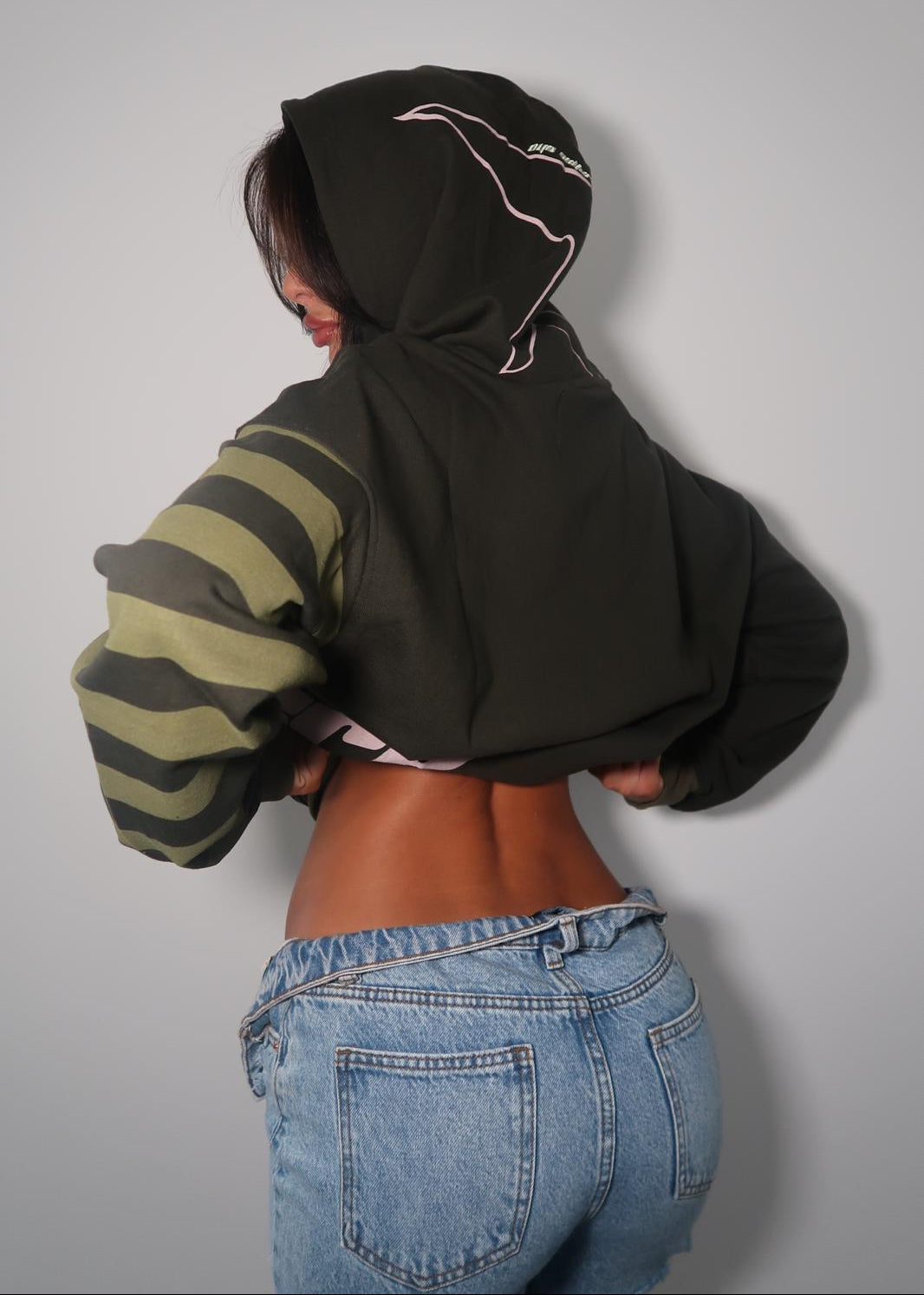 A person shown from behind wearing a cropped black hoodie with a light pink graphic outline on the hood and olive green and black striped sleeves, paired with blue jeans against a plain light gray background.