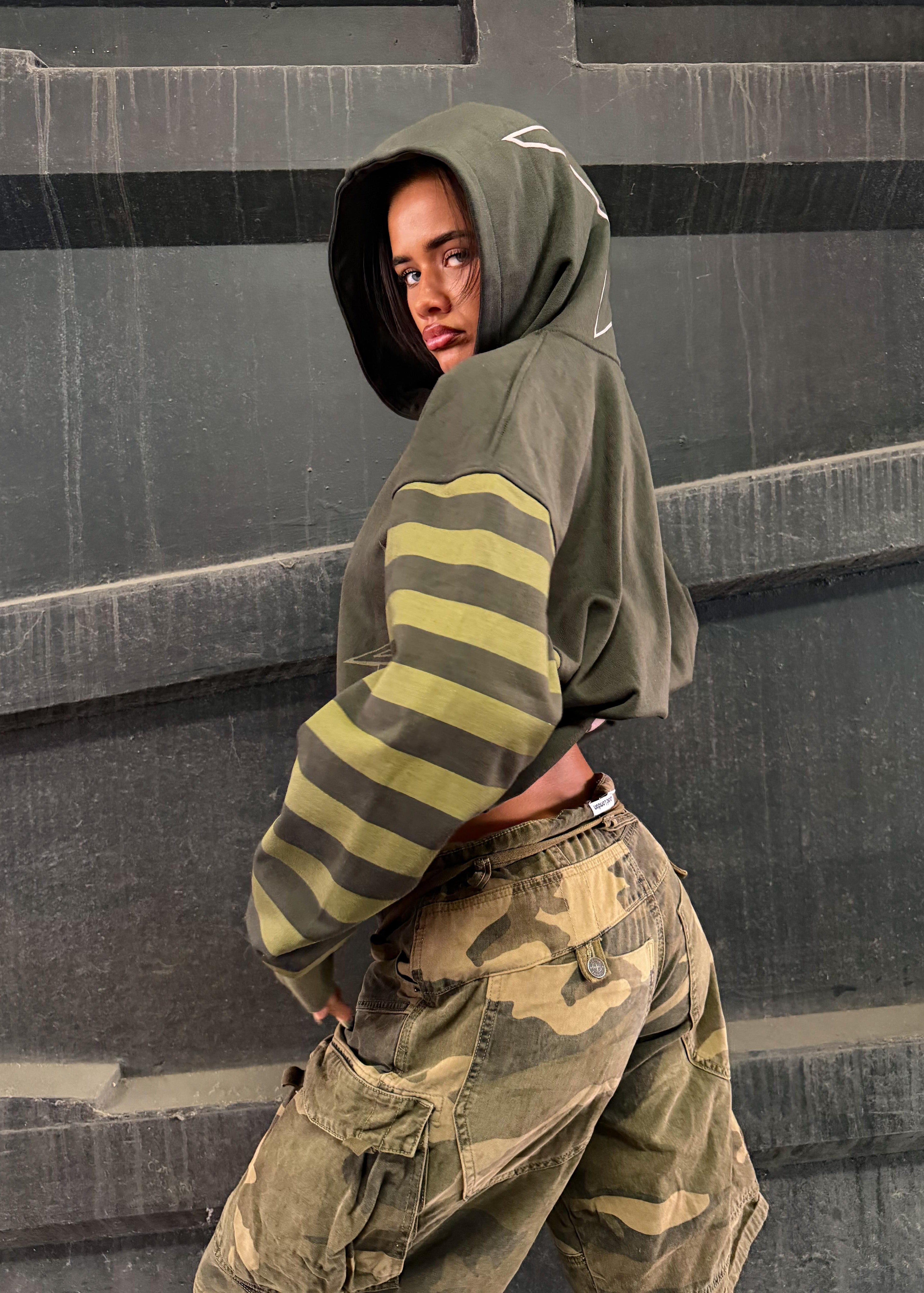 A model wearing a cropped olive green hoodie with yellow-green and dark gray horizontal stripes on the sleeves, paired with camouflage cargo pants, posing against a dark gray concrete wall.