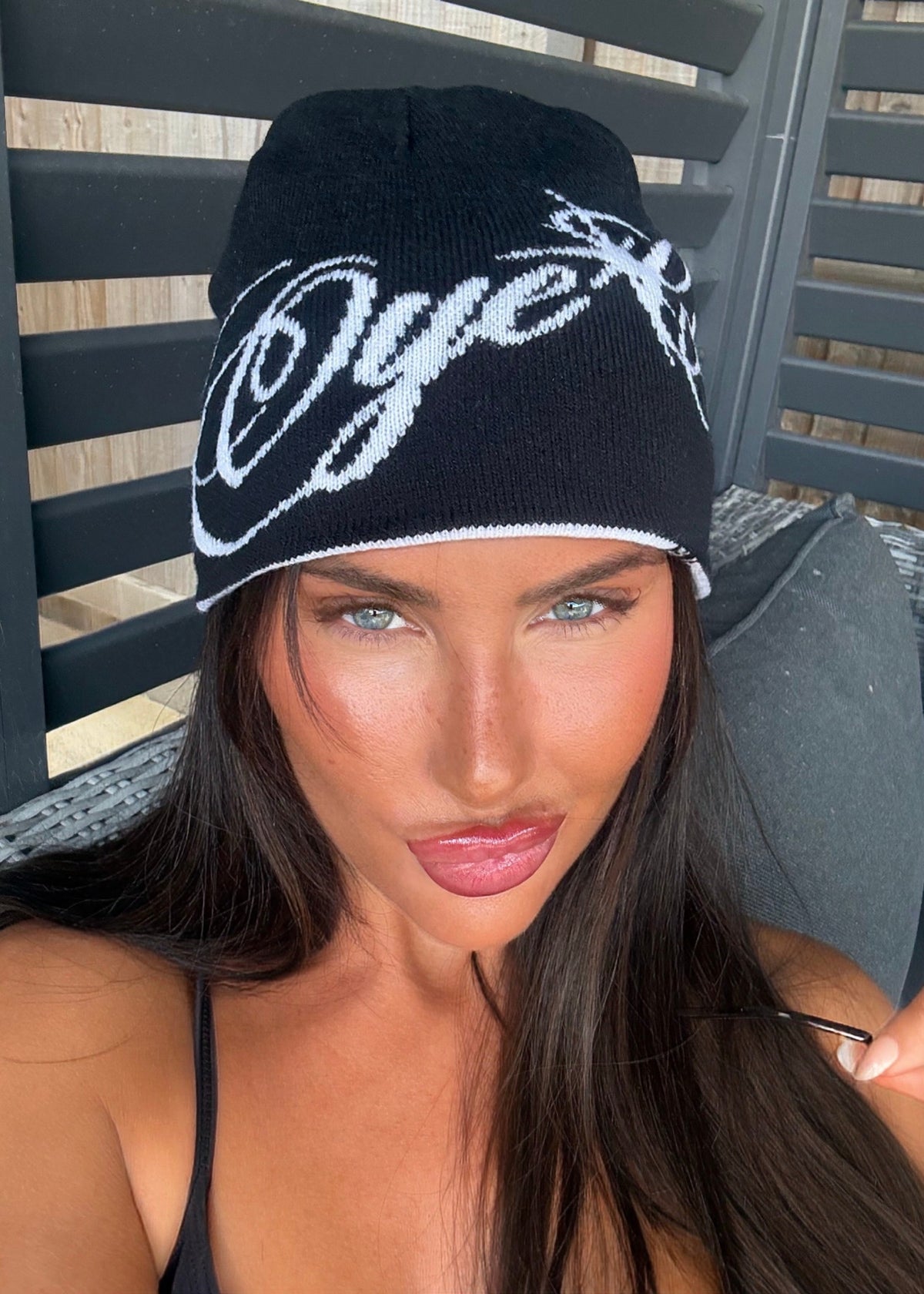 A woman wearing a black knit beanie with white embroidered script lettering, photographed in a close-up selfie-style shot against a dark slatted outdoor chair.