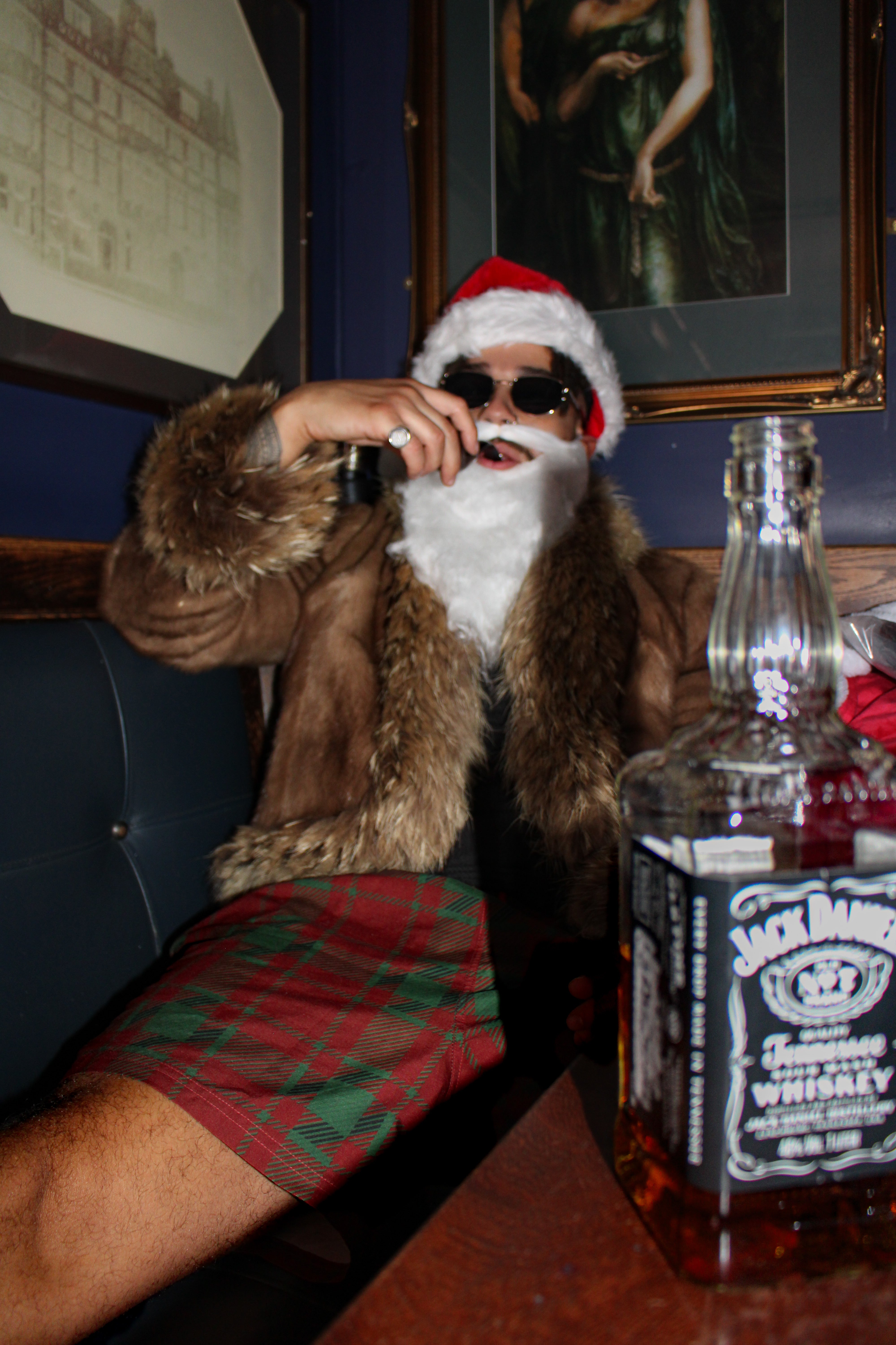 Person in Santa Claus outfit with fur coat and plaid skirt sitting at a bar with a Jack Daniel's bottle.