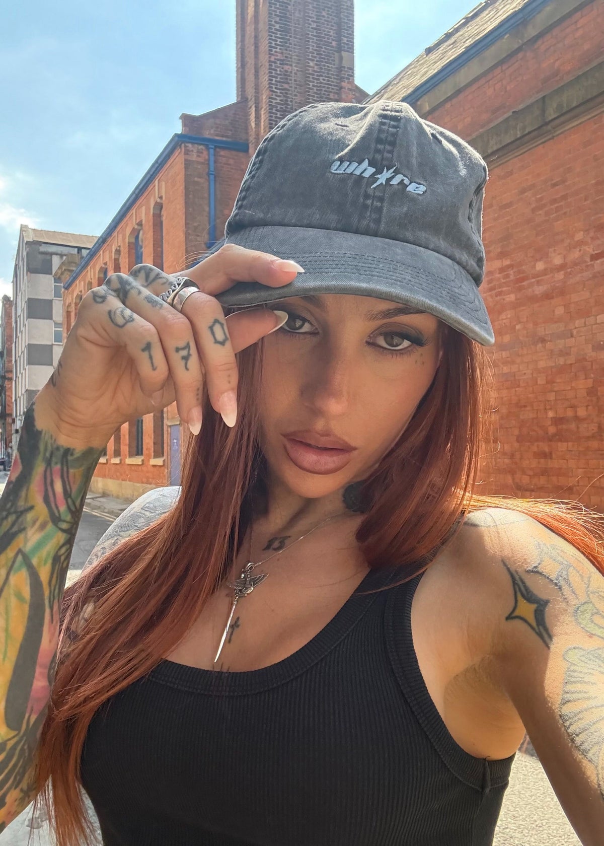 A woman with long red hair and visible tattoos wears a dark gray washed baseball cap with the word 'whya' embroidered on the front, posing outdoors in front of brick buildings under a blue sky.