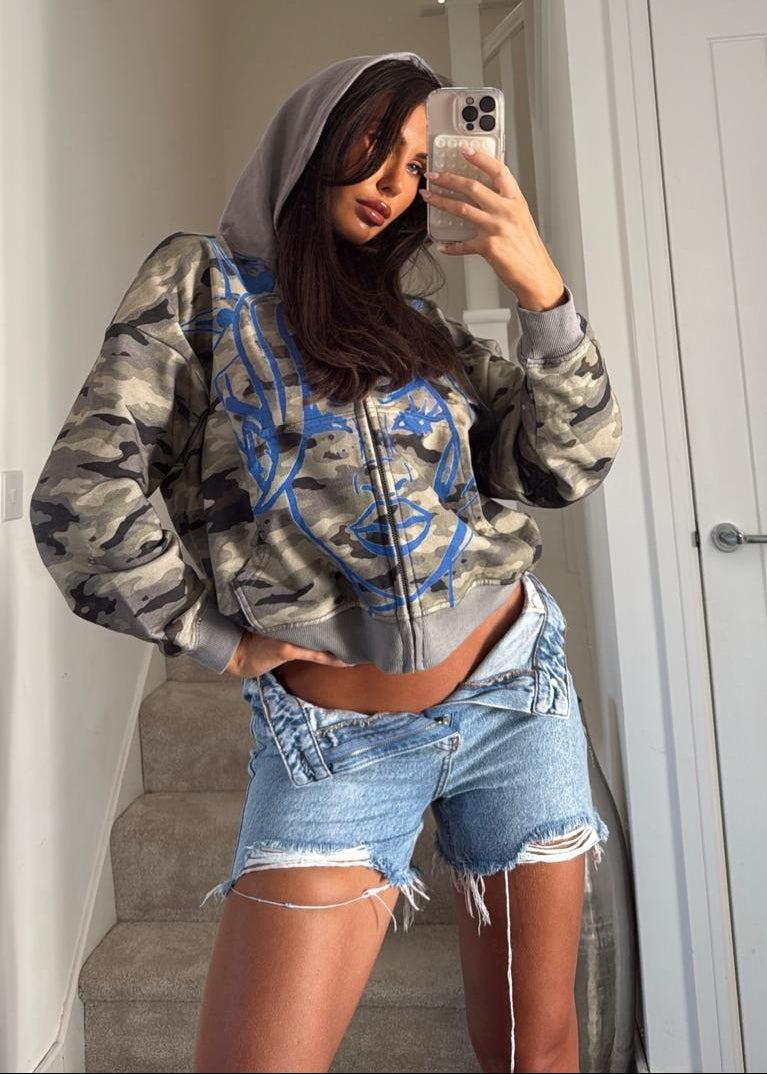 A woman takes a mirror selfie wearing a camouflage print zip-up hoodie with blue graphic details, a gray hood, and light-wash denim shorts in an indoor setting.