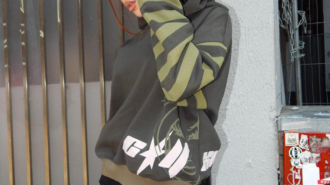 A woman with red hair and leg tattoos models a dark olive green and olive green striped hoodie with white graphic lettering, paired with black shorts, leaning against a gray wall next to a metal gate.