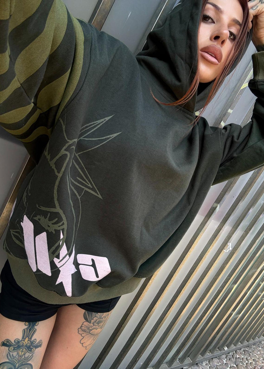 A person wearing an oversized dark olive green hoodie with a large light graphic print on the front, posed against a corrugated metal wall.