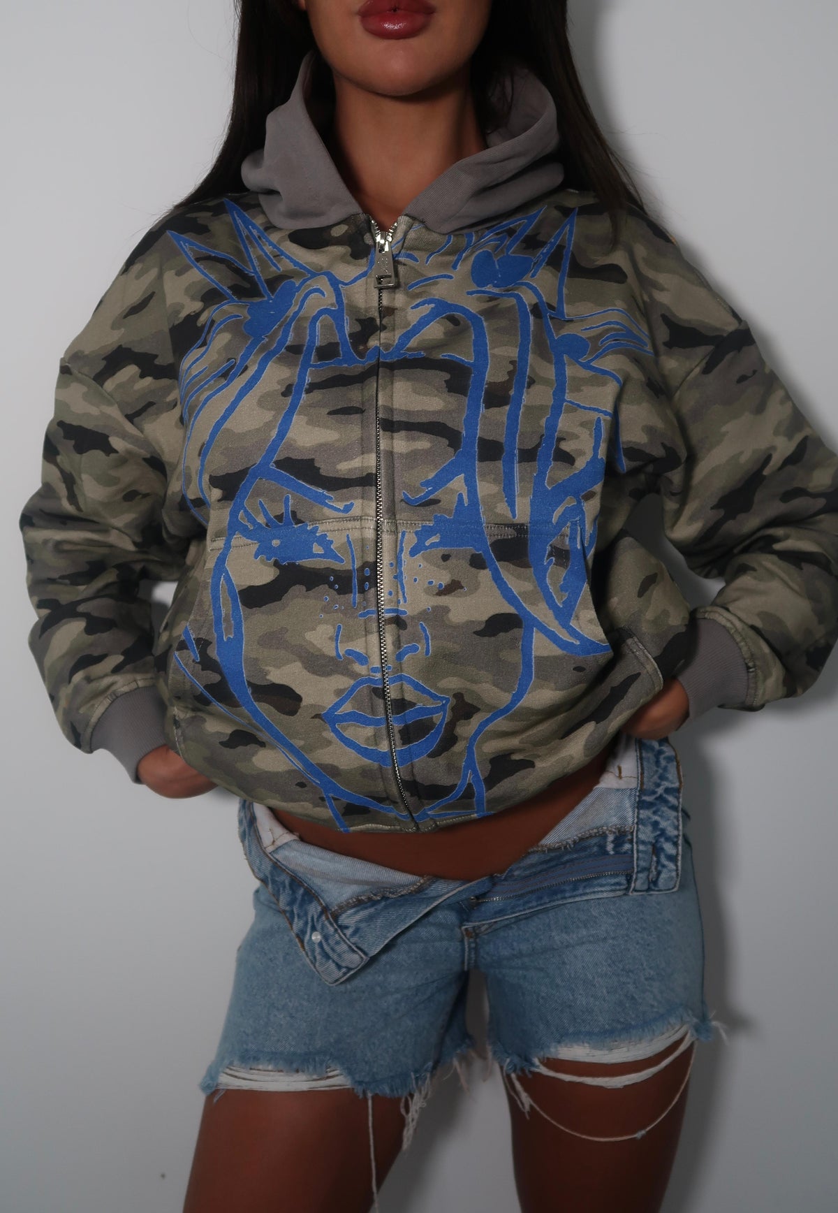 A woman wearing a cropped zip-up hoodie with a green and brown camouflage pattern and a large blue graphic of a face on the front, paired with distressed denim shorts.