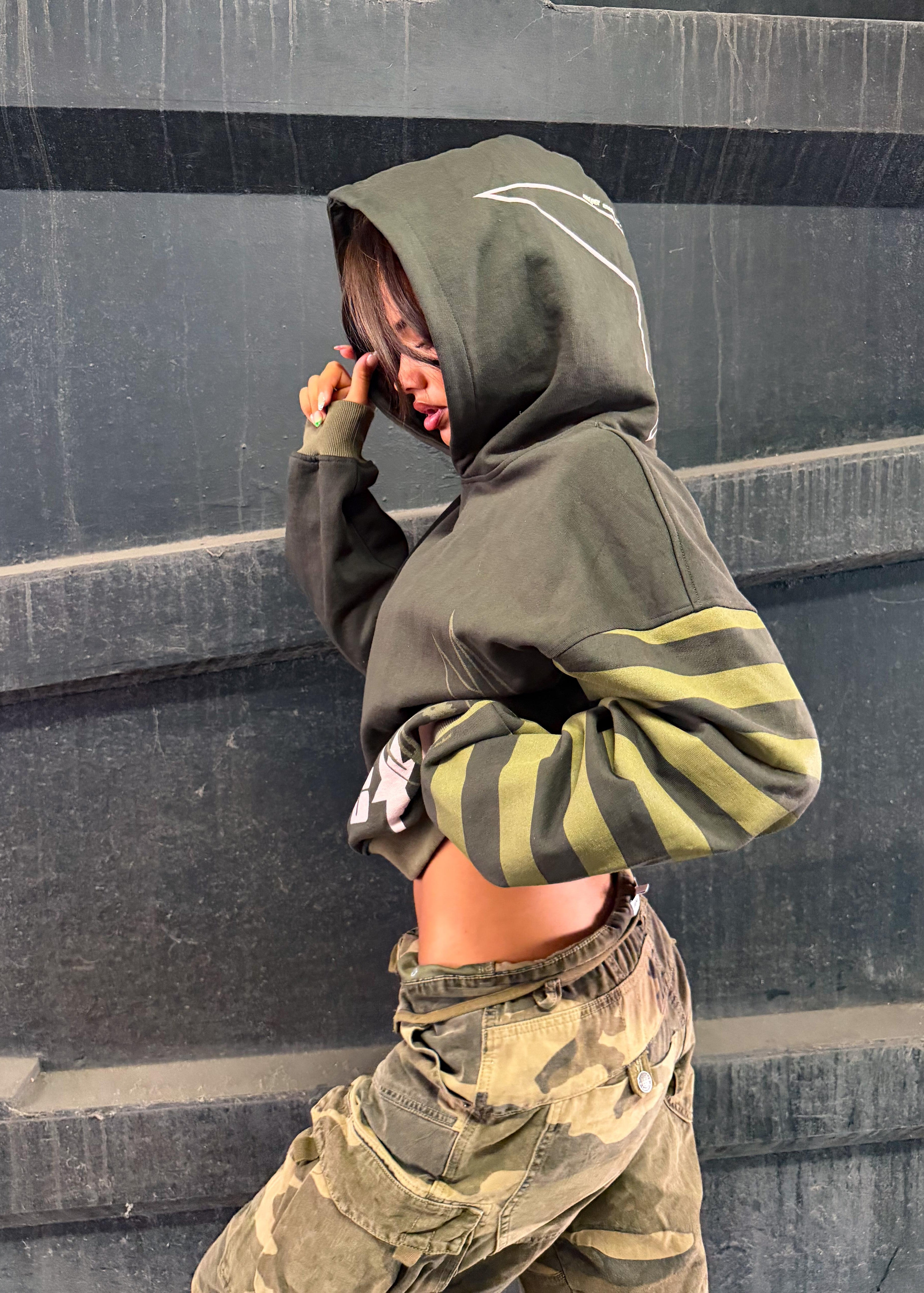 A person wearing a cropped olive green hoodie with yellow-green striped sleeves and camouflage cargo pants, posing against a dark gray concrete wall.