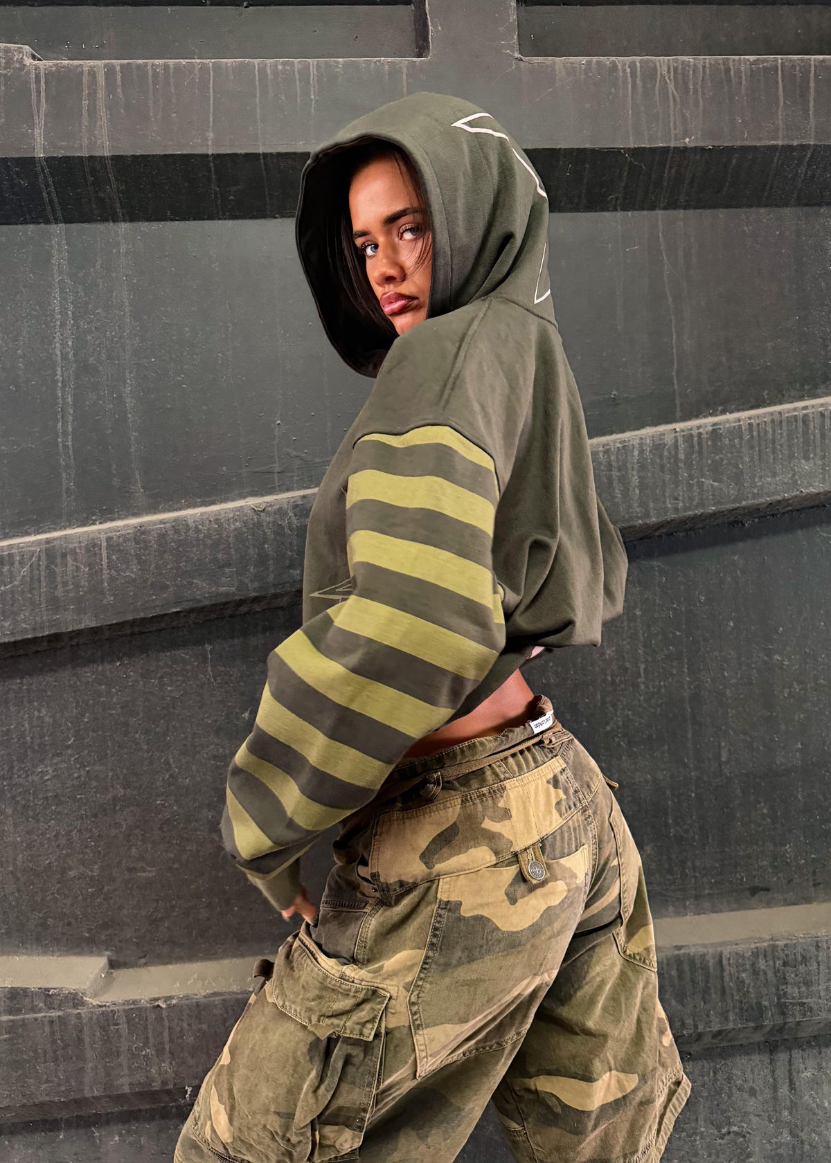 A model wearing a cropped olive green hoodie with yellow-green and dark gray horizontal stripes on the sleeves, paired with camouflage cargo pants, posing against a dark gray concrete wall.